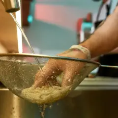 Ręce w rękawiczkach płuczą makaron pod bieżącą wodą, by zapobiec jego sklejaniu.