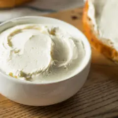 Biały serek kremowy w miseczce obok kromki chleba z serkiem.
