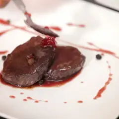 Elegancko podany comber z jelenia, polany sosem z owoców leśnych, ozdobiony ziarnami pieprzu i jagodami.