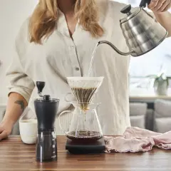 Kobieta parzy kawę metodą przelewową. Na blacie stoi młynek, kubek i dripper z papierowym filtrem do kawy rozmiar 2.