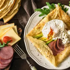 Złożone crepes z jajkiem w koszulce, awokado, szynką i serem. Obok stos naleśników, pomidorki i rukola.
