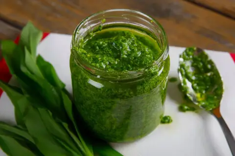 Słoik pełen aromatycznego pesto z czosnku niedźwiedziego, obok świeże liście i łyżka z porcją zielonego sosu.