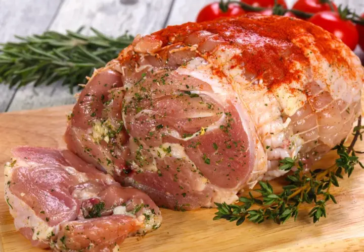 Surowa szynka z kurczaka, doprawiona ziołami i papryką, gotowa do pieczenia. Obok gałązki rozmarynu i tymianku, w tle pomidorki koktajlowe.