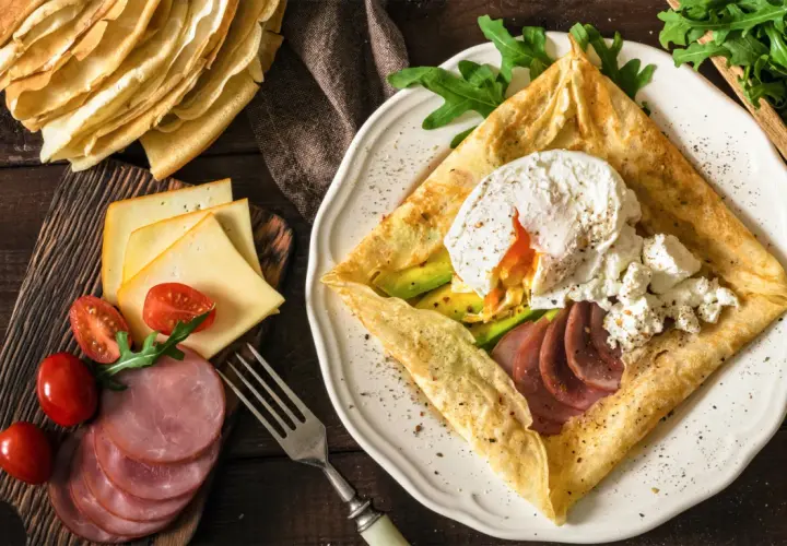 Złożone crepes z jajkiem w koszulce, awokado, szynką i serem. Obok stos naleśników, pomidorki i rukola.