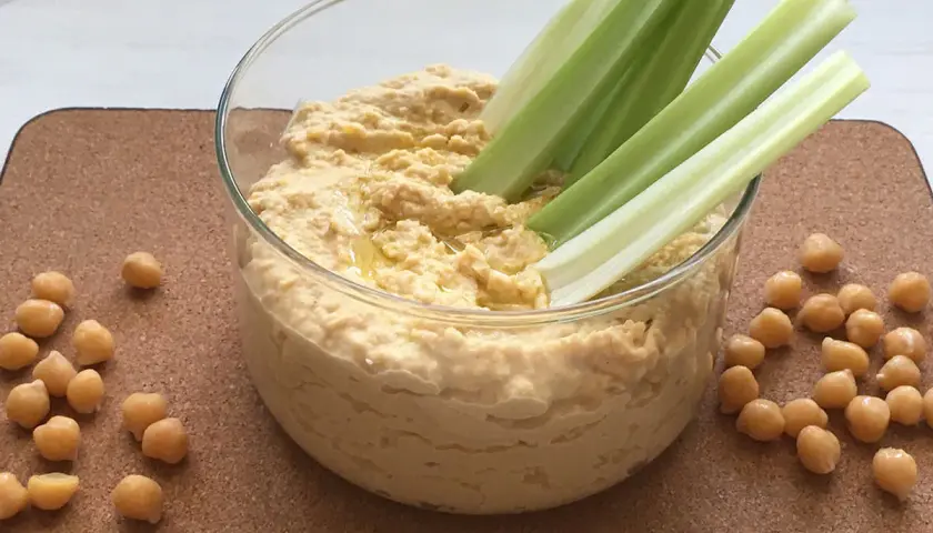Domowy hummus przepis: kremowa pasta z ciecierzycy w szklanej misce, podana z łodygami selera i ziarnami ciecierzycy.