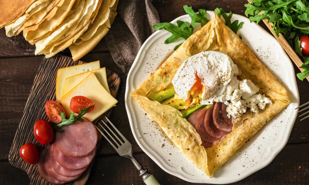 Złożone crepes z jajkiem w koszulce, awokado, szynką i serem. Obok stos naleśników, pomidorki i rukola.
