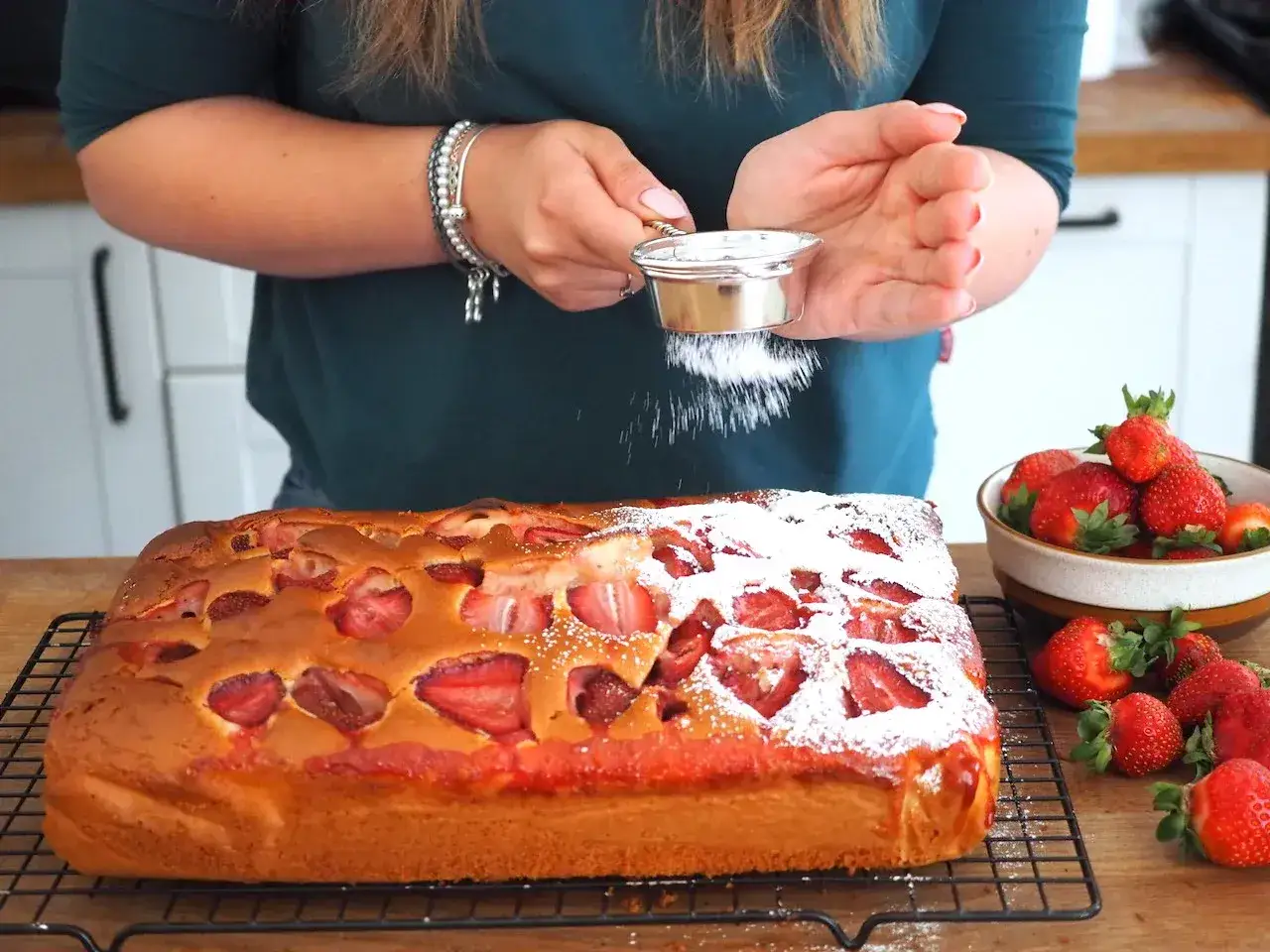 Puszyste ciasto na kefirze z truskawkami, posypywane cukrem pudrem. Obok miseczka świeżych owoców.