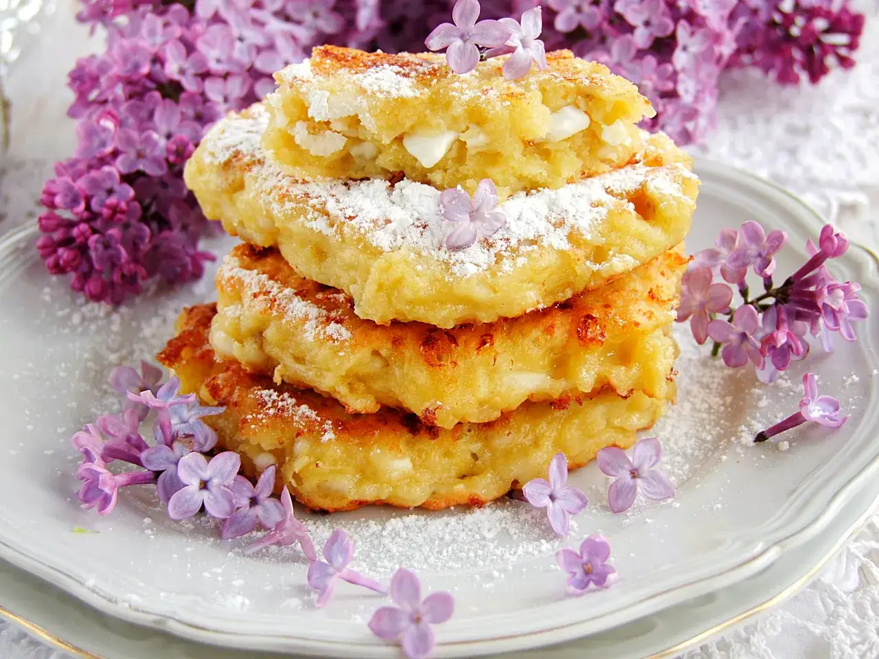 Stos placuszków z serka wiejskiego i banana, posypanych cukrem pudrem, ozdobiony kwitnącym bzem.