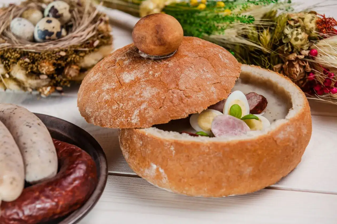 Smaczne obiady domowe przepisy: żurek w chlebie z kiełbasą i jajkiem, obok biała kiełbasa i jajka przepiórcze w gnieździe.