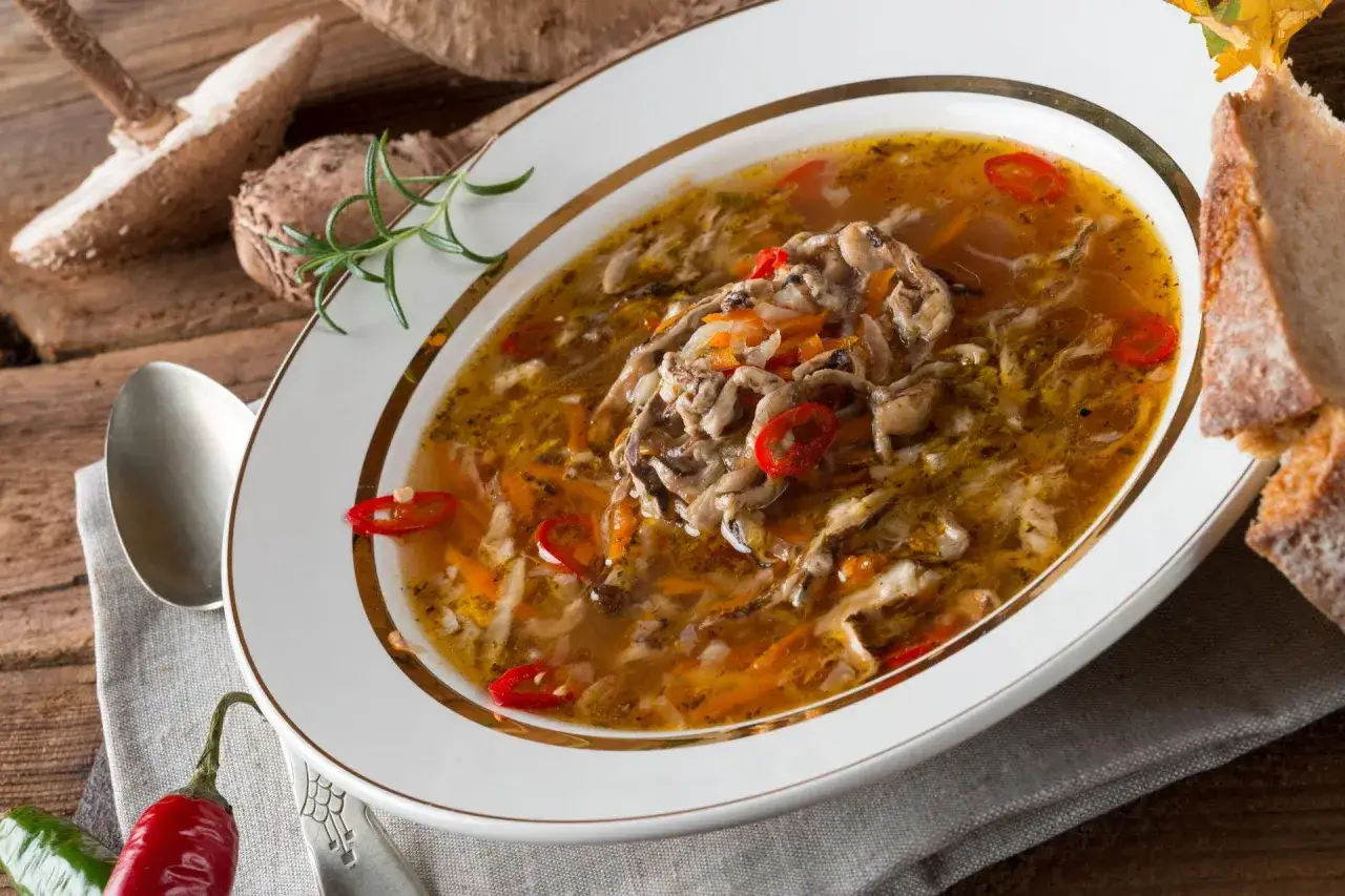 Aromaticzne flaczki z kani, doprawione papryczką chili, podane w białej misce z złotym rantem, obok kromka chleba i grzyby.