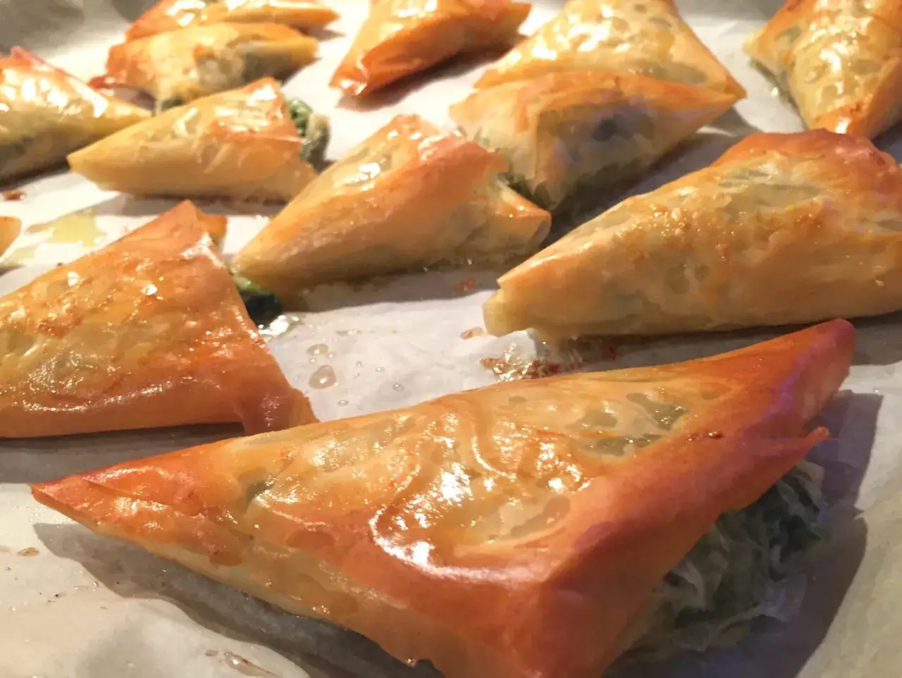 Złociste, chrupiące trójkąciki spanakopita z nadzieniem szpinakowo-serowym, pieczone na papierze do pieczenia.